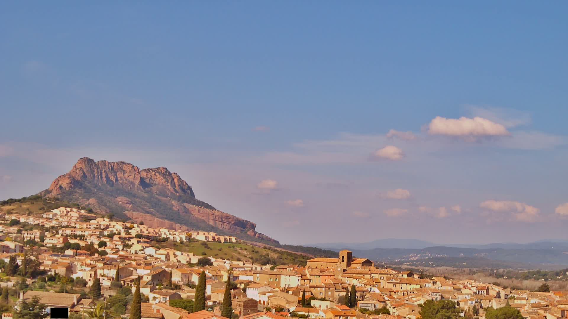 A town of tan-colored buildings nestled against a large, reddish-brown mountain under a mostly clear, pale blue sky with a few puffy white clouds.