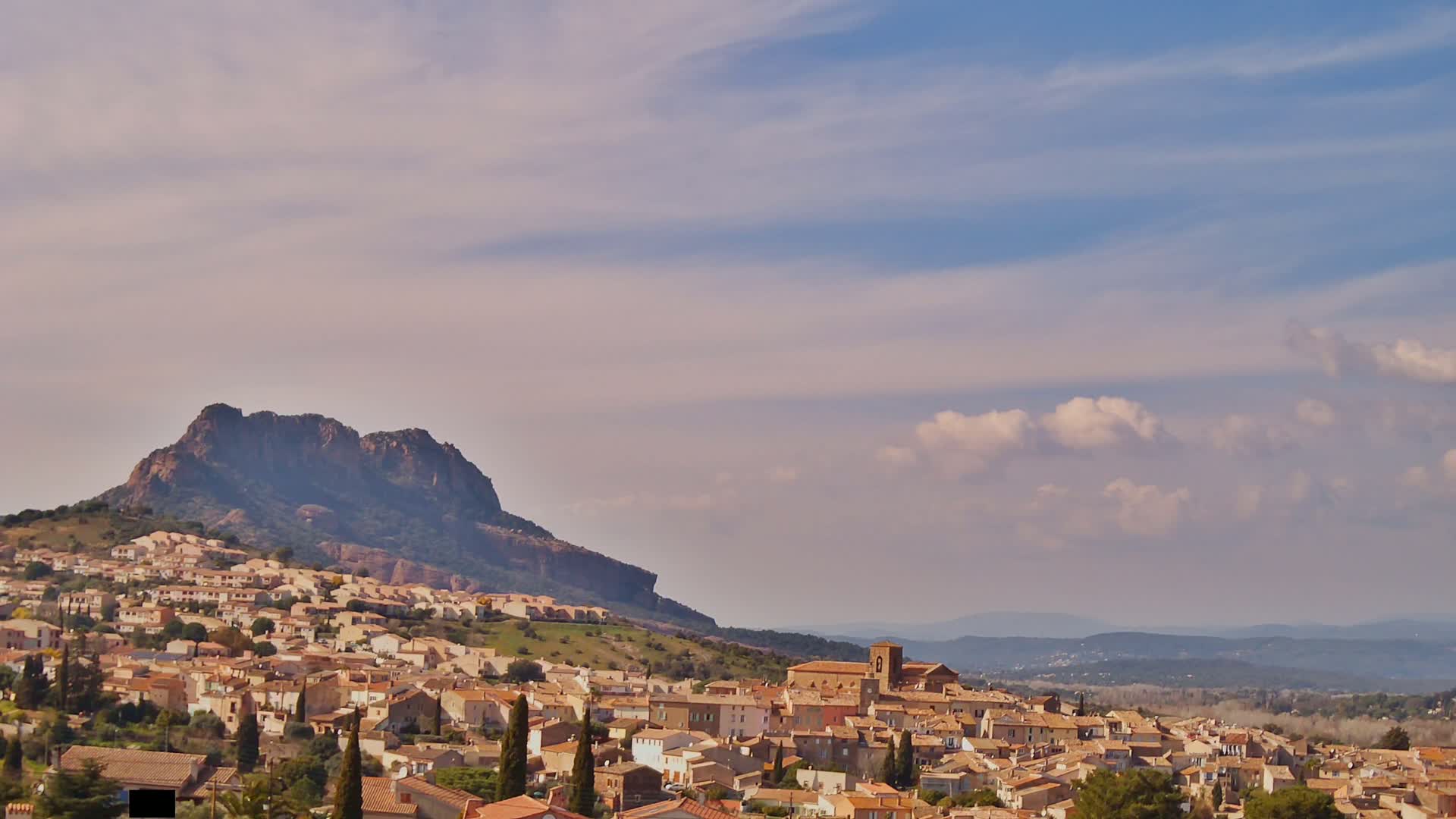 A small town of tan and brown buildings sits on a hillside beneath a large, dark mountain under a mostly clear, pale sky with a few puffy clouds.