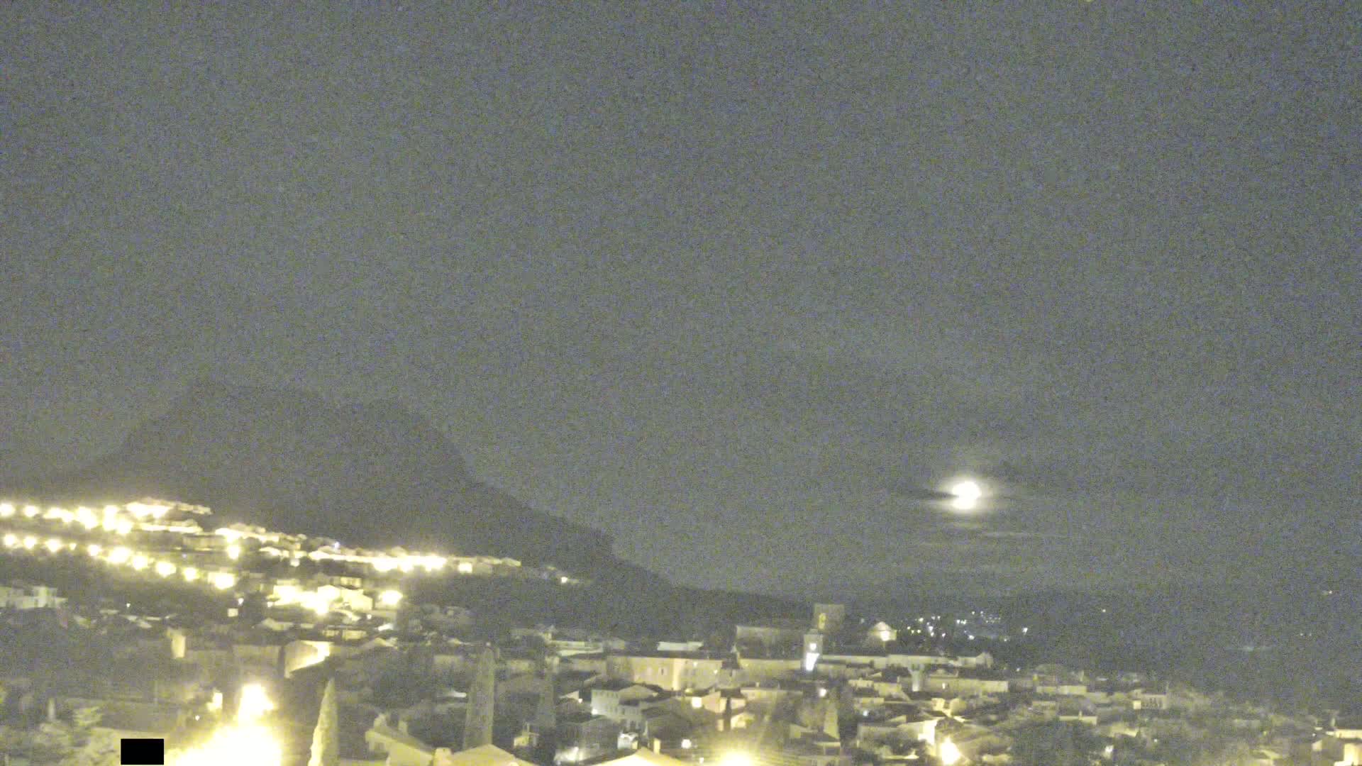 A nighttime view of a town nestled against a hill, illuminated by lights, under a mostly clear sky with a bright moon.