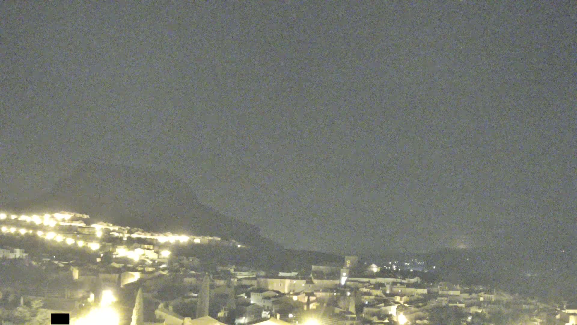 A nighttime view of a small town nestled against a hill under a cloudy sky.