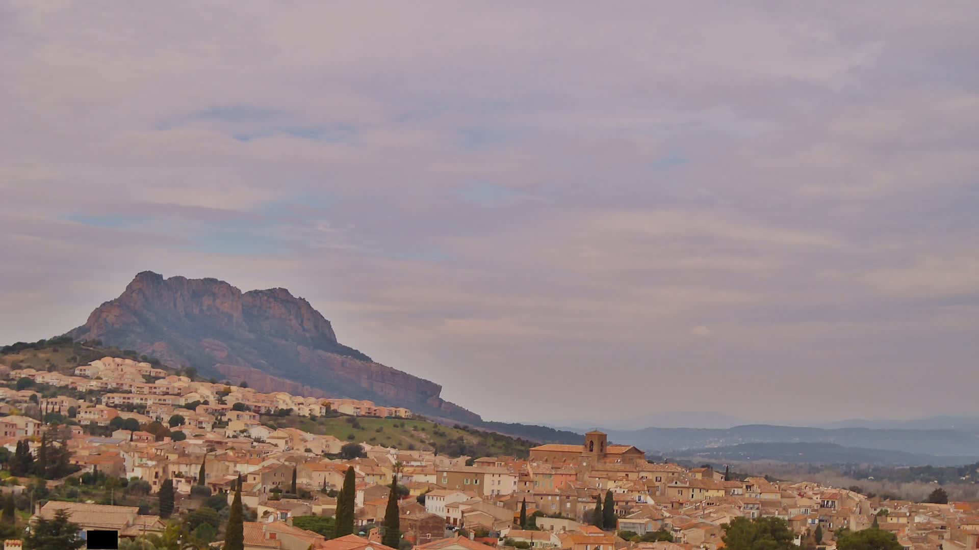 A hilltop town of tan buildings nestles against a large, dark mountain under a pale, cloudy sky.