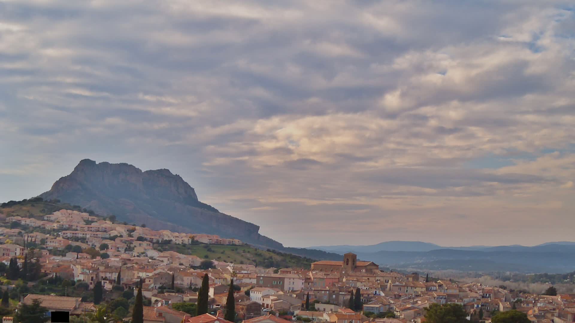 A sprawling town of tan-colored buildings nestles at the foot of a large, dark mountain under a mostly cloudy sky.