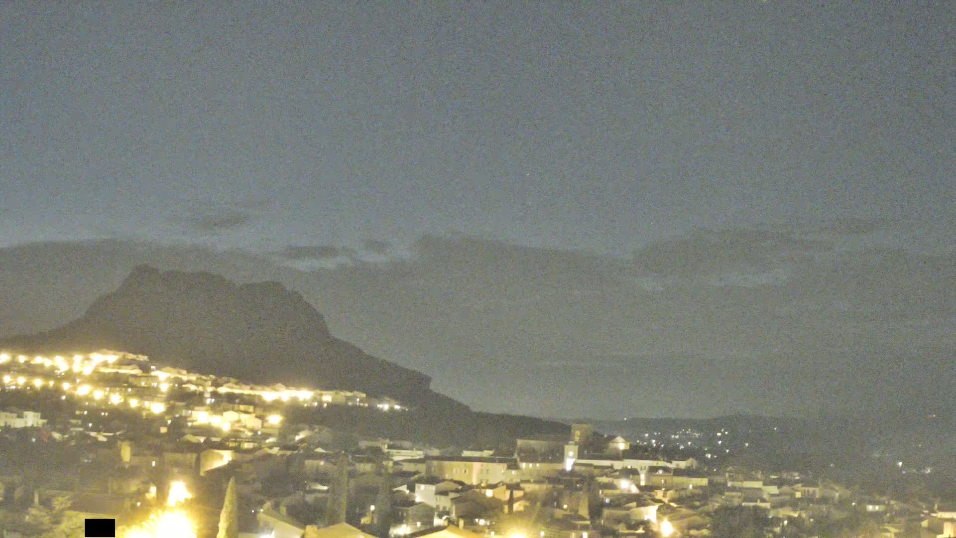 A nighttime view of a town nestled at the base of a large, dark mountain under a mostly clear, dusky sky.