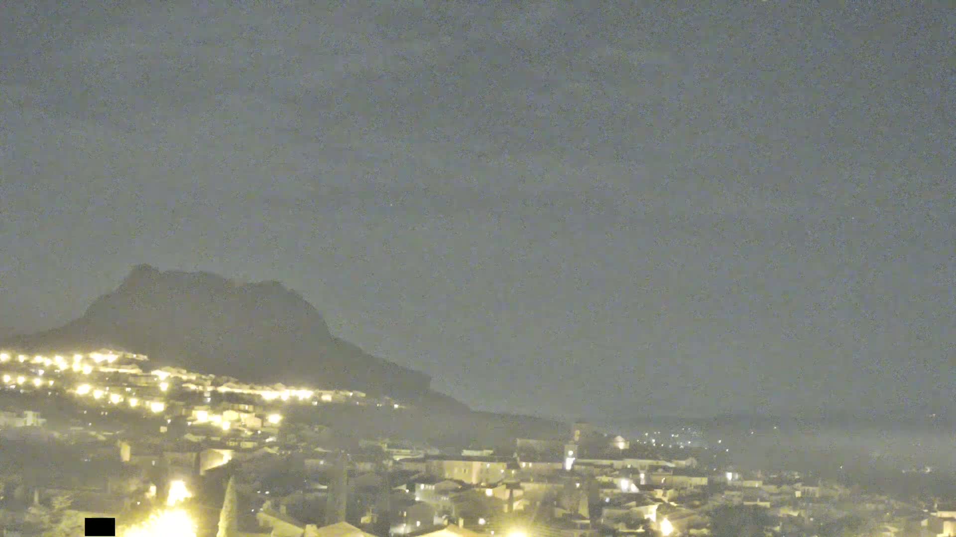 A nighttime view shows a town illuminated by lights nestled below a dark, large hill under a hazy sky.
