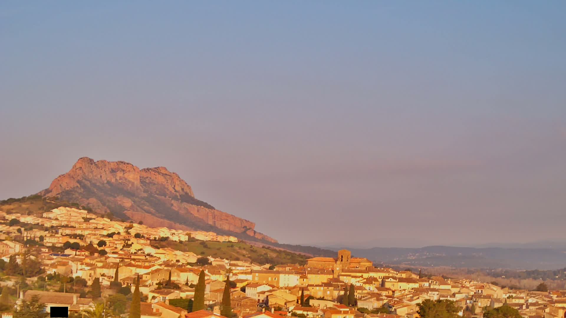 A small town of tan and yellow buildings nestles at the base of a large, reddish-brown mountain under a pale, clear sky.