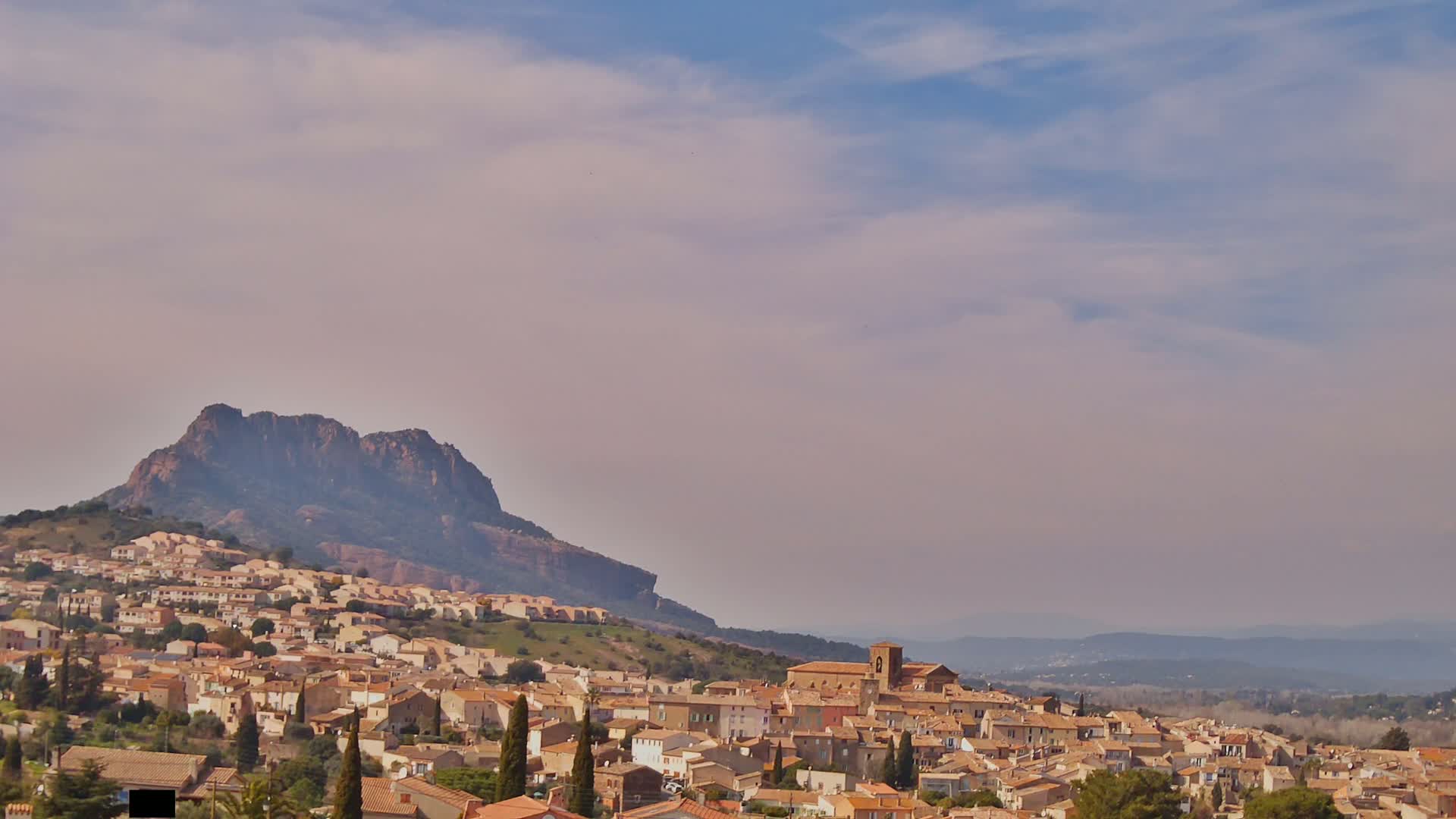 A tan-colored village nestled against a large, dark mountain under a hazy, partly cloudy sky.