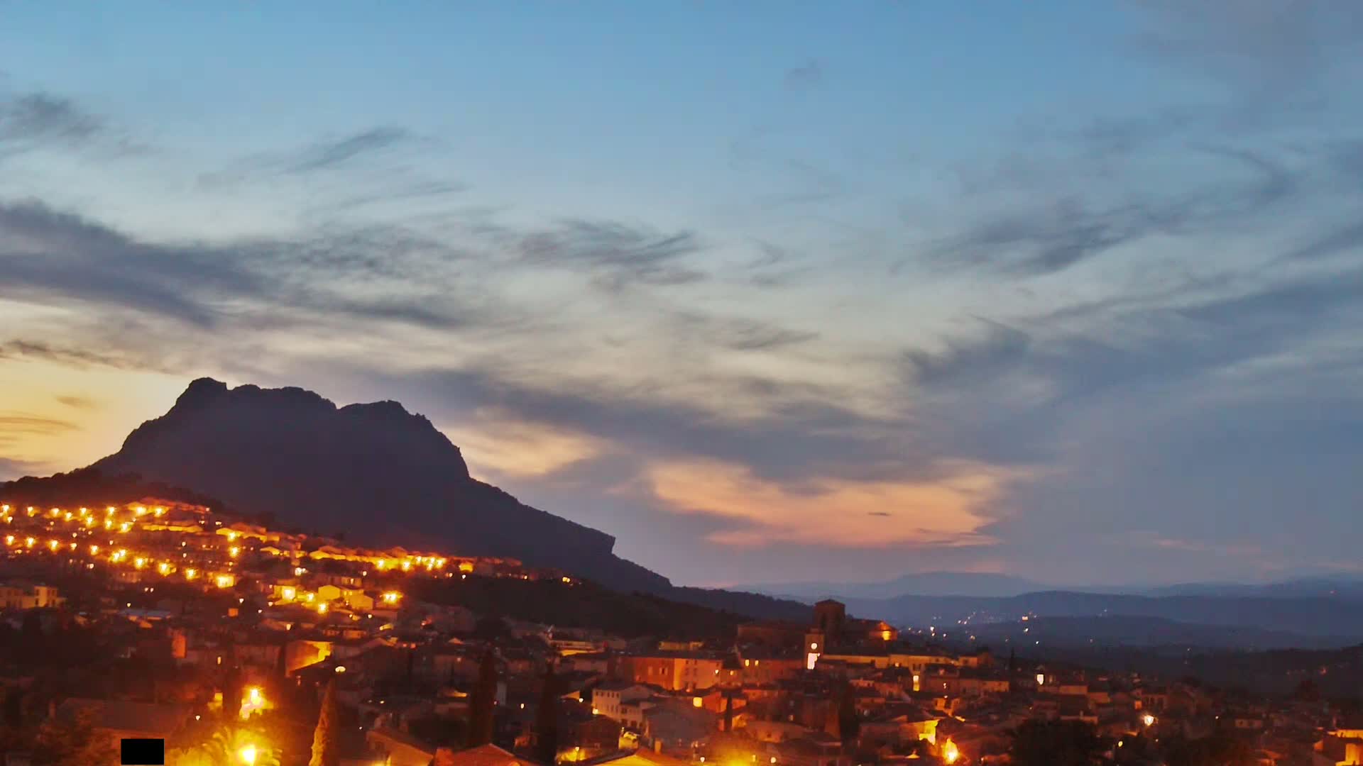 A village with illuminated buildings nestles at the foot of a large, dark mountain under a twilight sky with a mix of clouds.