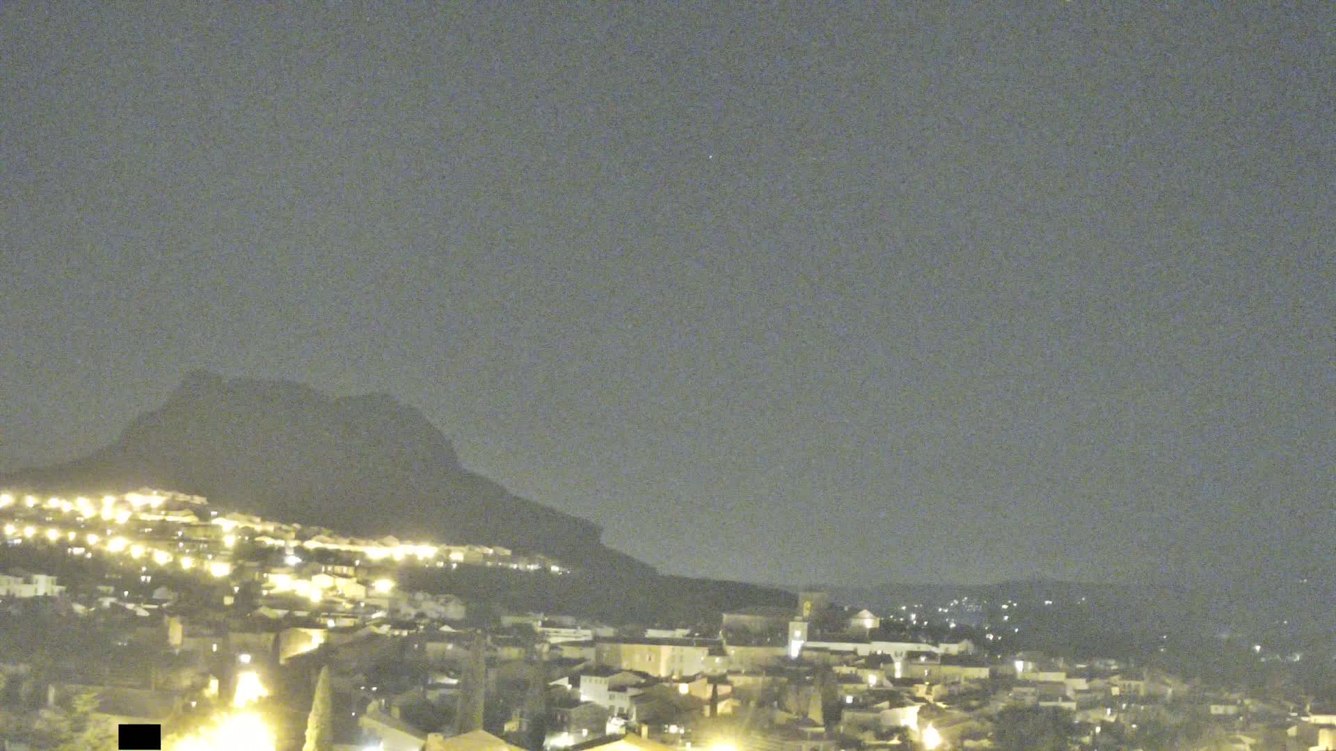 A nighttime view of a town illuminated by lights, nestled against a dark mountain under a clear, dusky sky.