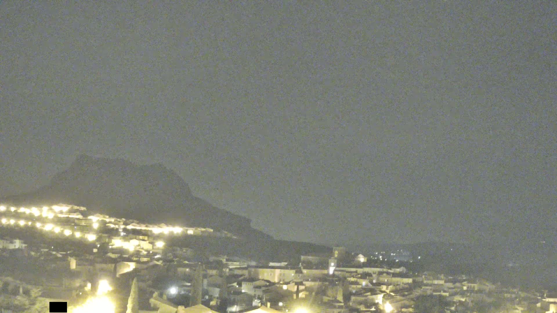 A nighttime view of a town nestled at the base of a large hill under a cloudy sky.