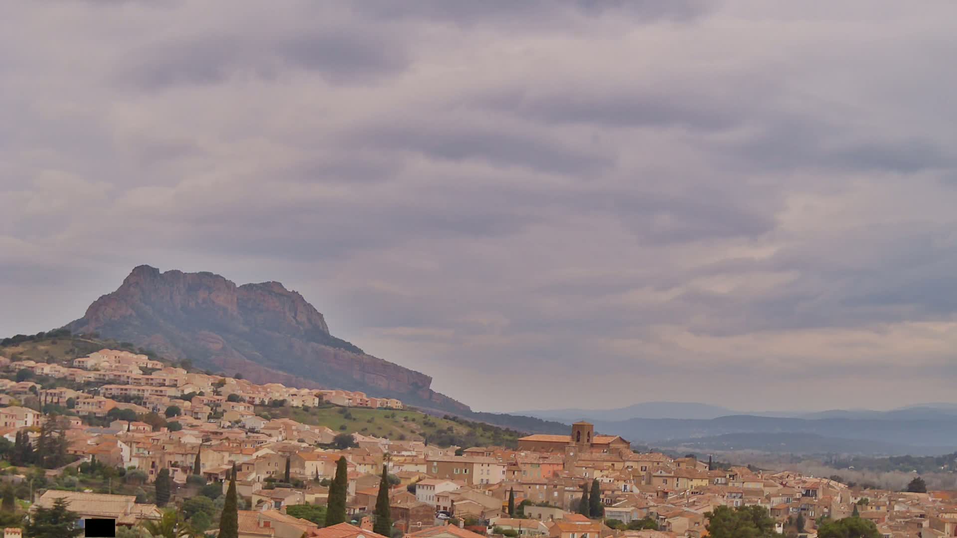 A hilltop town with tan buildings stretches across a hillside beneath a large, rocky mountain under a cloudy sky.