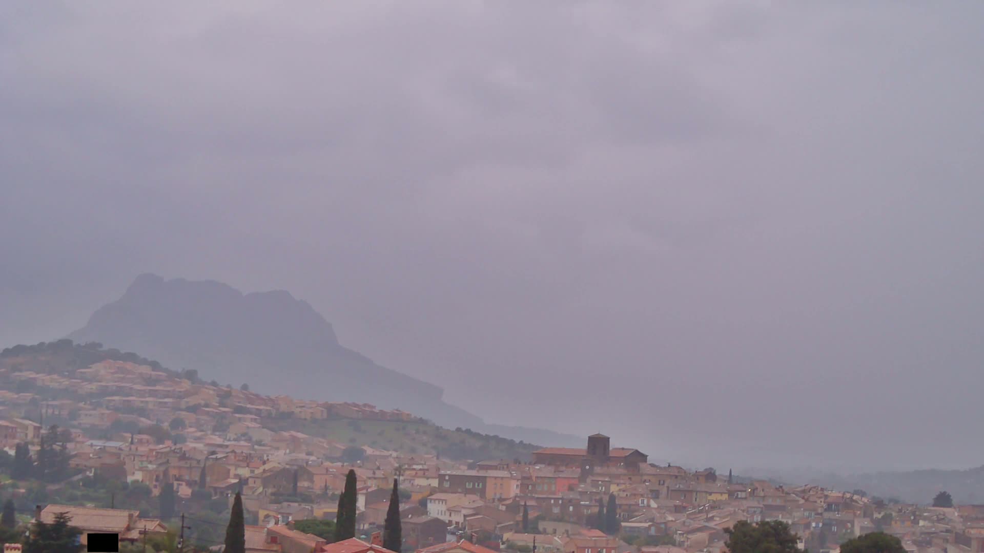 A hazy, overcast sky hangs over a hillside town nestled against a large, dark mountain.