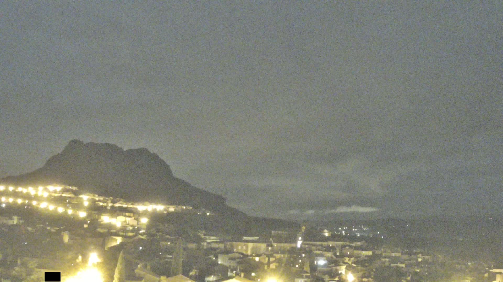 A nighttime view of a town nestled at the base of a dark mountain under a cloudy, overcast sky.
