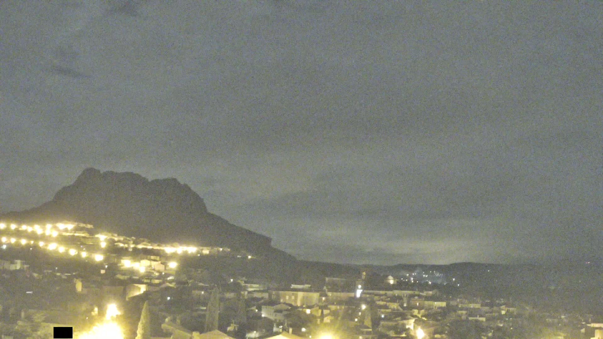 A nighttime view of a town illuminated by streetlights, nestled at the base of a dark mountain under a cloudy sky.