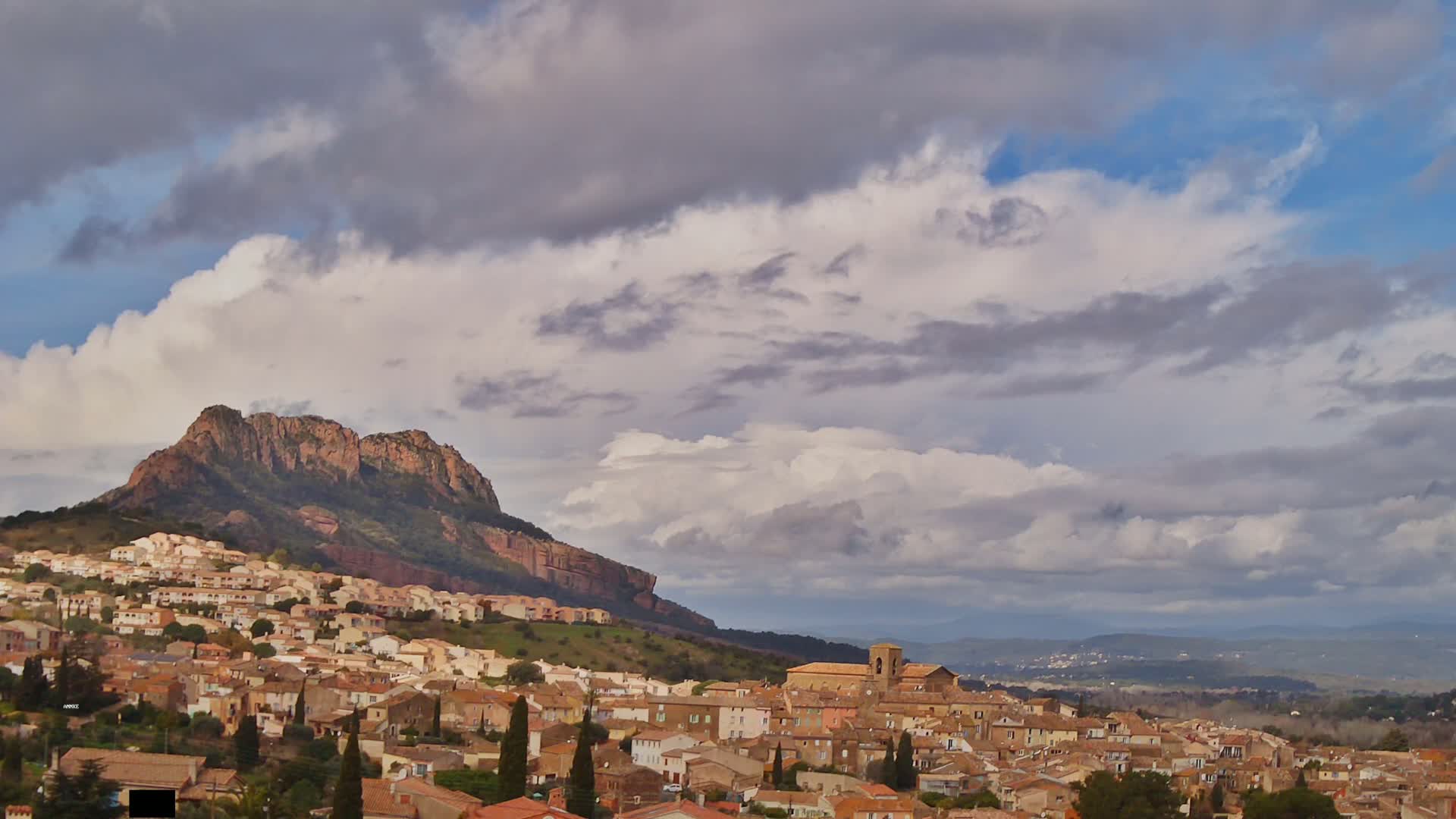 A sprawling town with terracotta rooftops nestles against a rocky hill under a mostly cloudy sky.