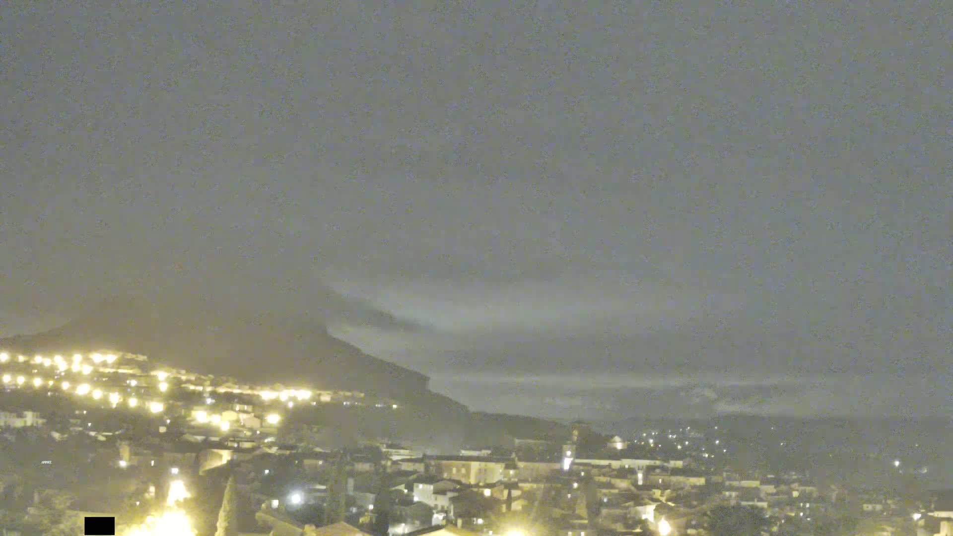 A nighttime view of a town nestled at the base of a mountain shrouded in fog under a cloudy sky.