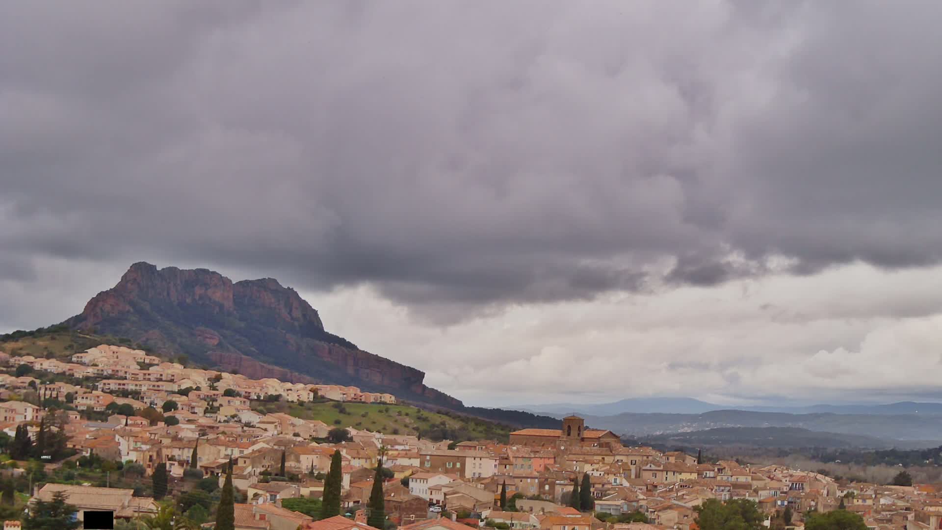 A sprawling town of tan and brown buildings nestles at the base of a large, dark-colored mountain under a cloudy, overcast sky.