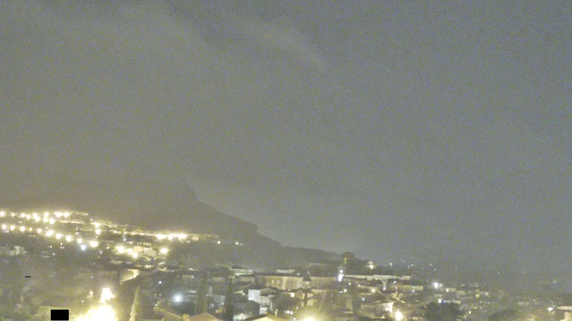 A night-time, hazy view of a town nestled at the base of a hill, with scattered lights illuminating the buildings below.