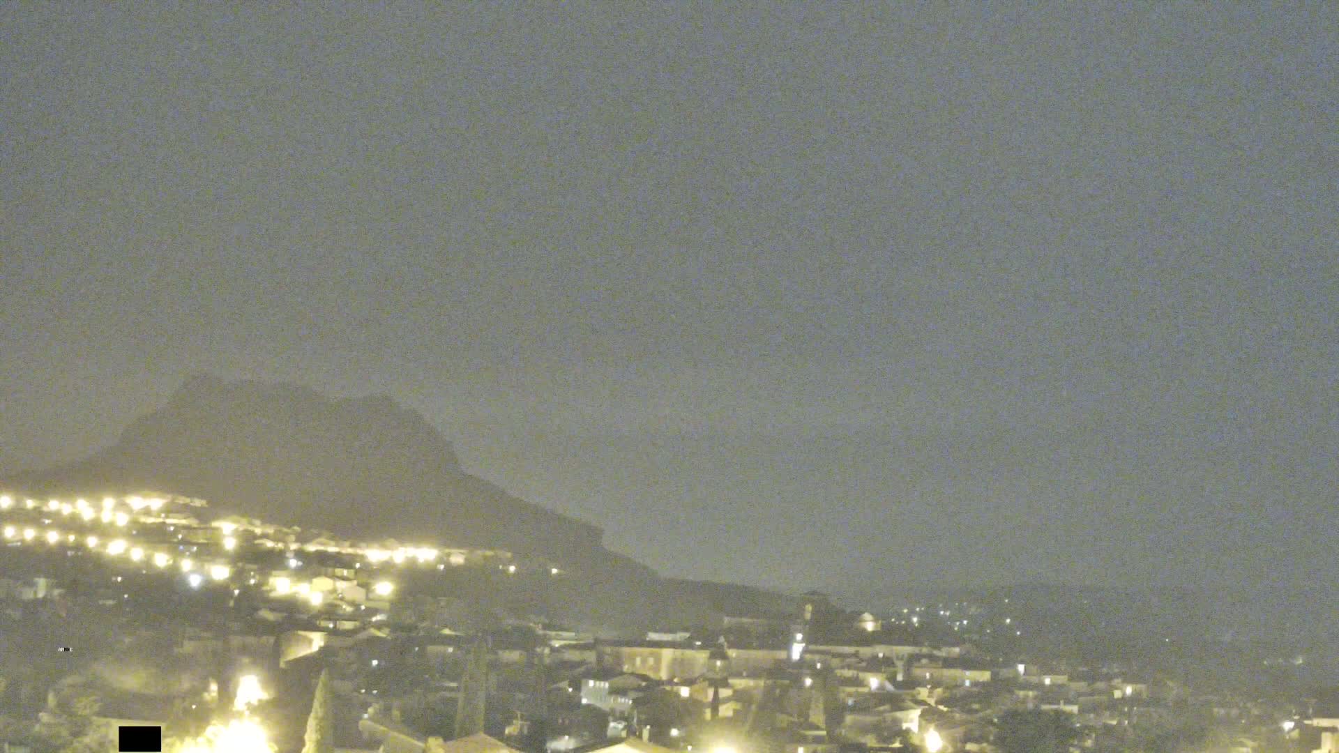 A hazy night scene shows a town nestled at the base of a dark mountain, with scattered lights illuminating the buildings below.