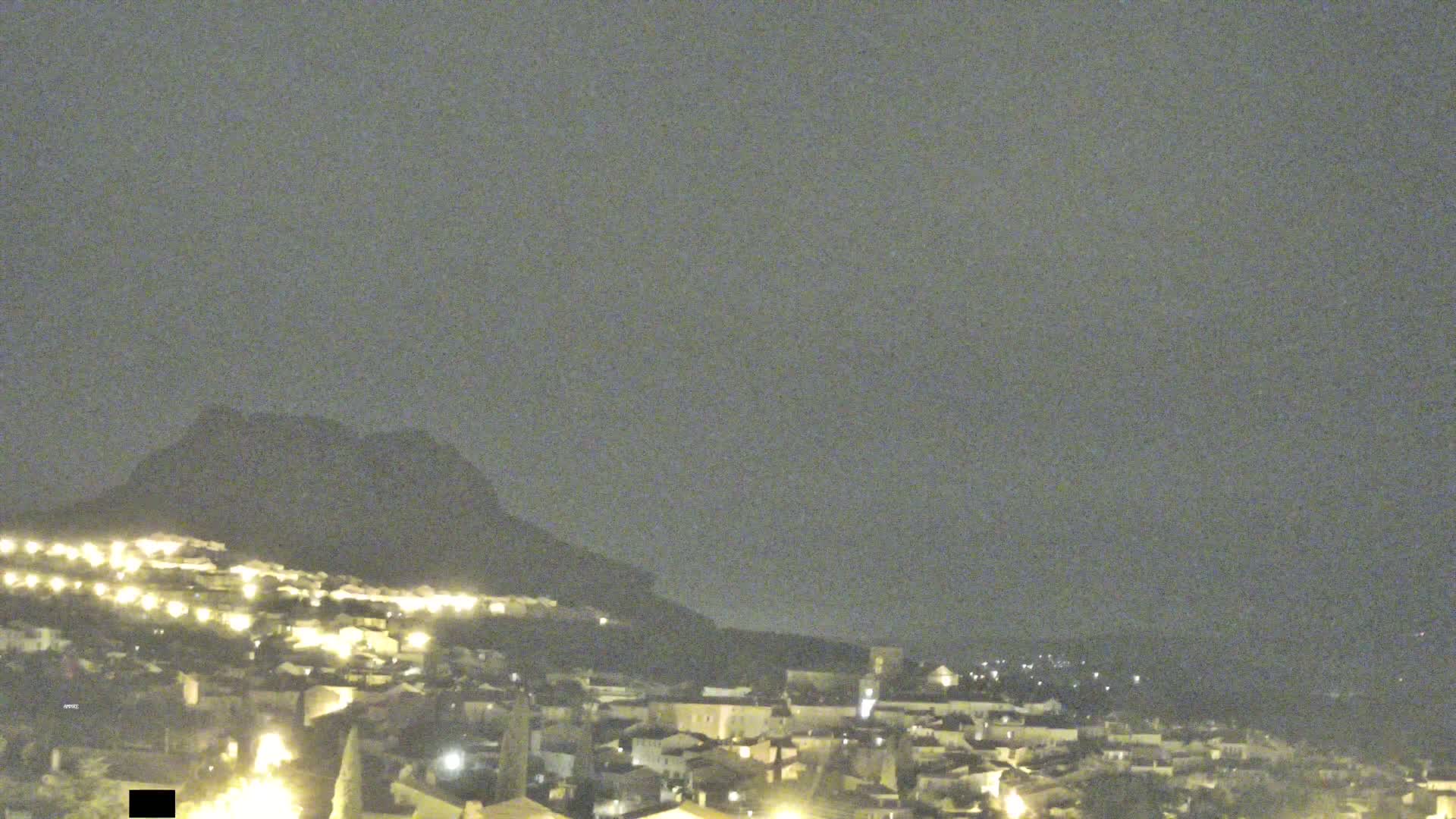 A nighttime view of a town illuminated by streetlights nestled against a dark mountain under a cloudy sky.