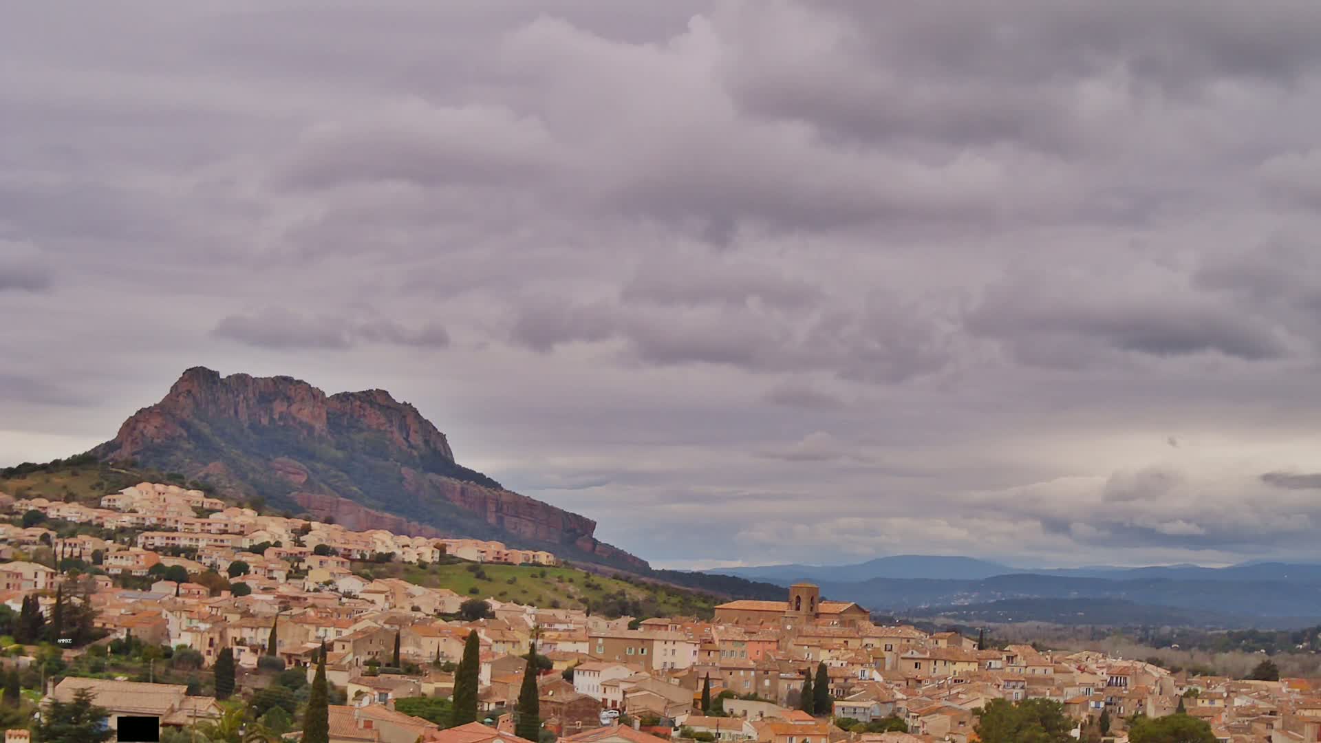 A hillside town of tan buildings nestles below a large, dark-red rock formation under a cloudy sky.