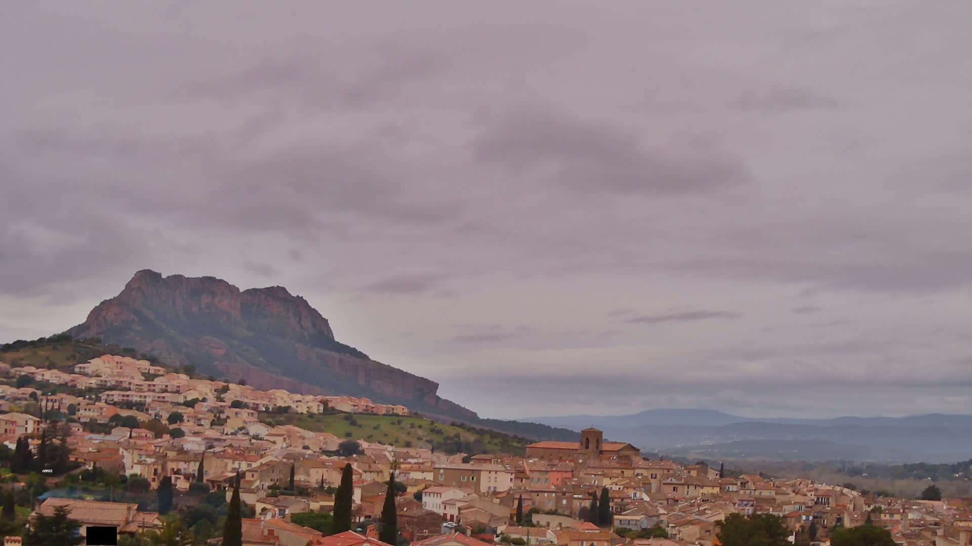 A hillside town with tan and brown buildings sits beneath a large, dark mountain under an overcast sky.