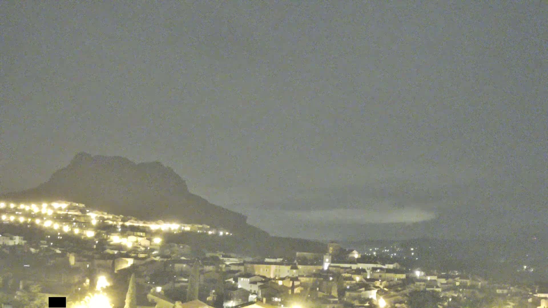 A nighttime view of a town nestled at the base of a dark mountain under a cloudy sky.