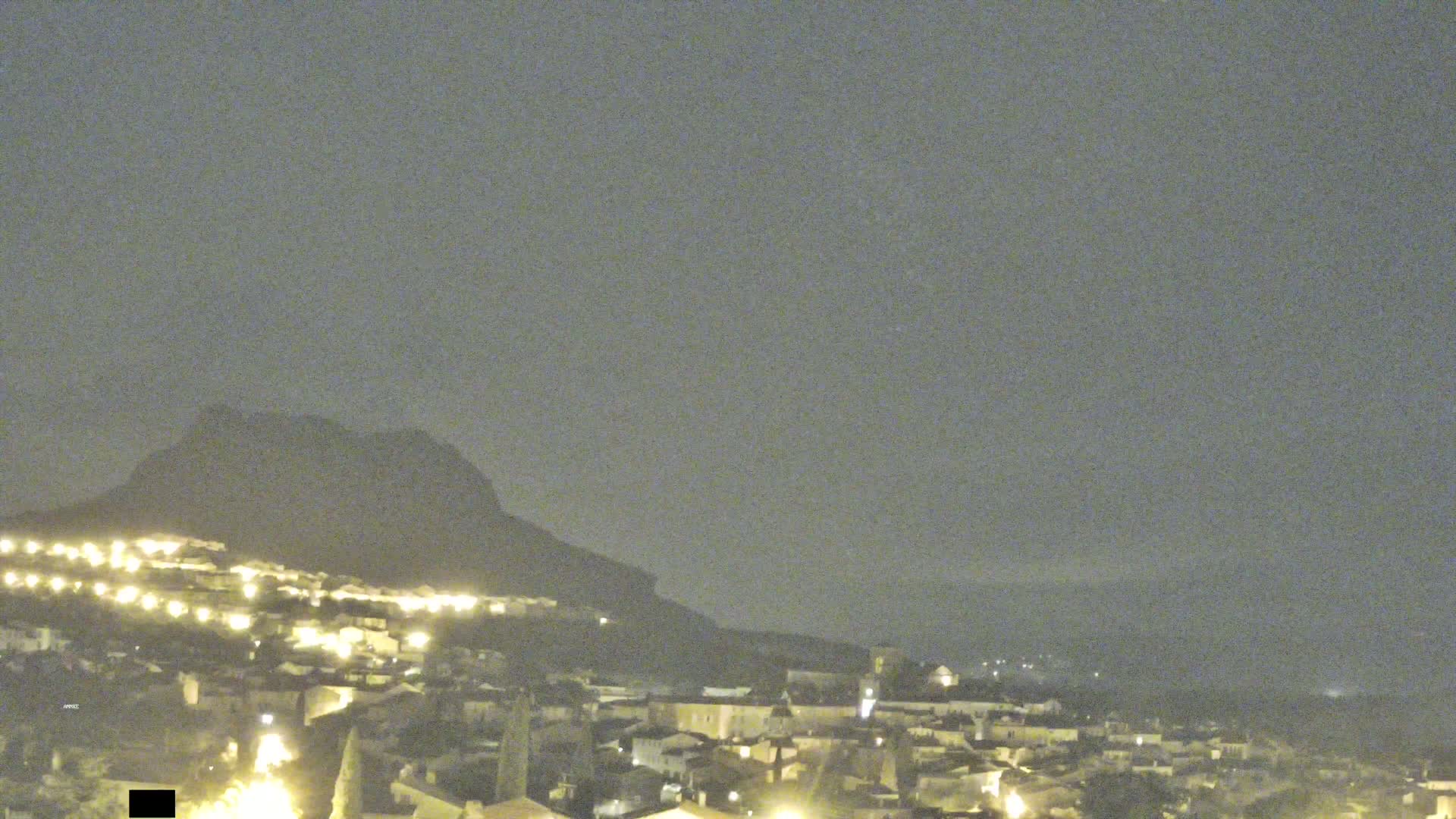 A nighttime view of a small town nestled at the base of a large, dark mountain under a hazy sky.