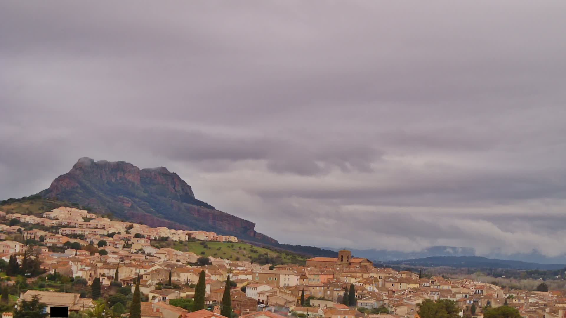 A hillside town of tan buildings nestles below a large, dark mountain under a mostly cloudy, overcast sky.