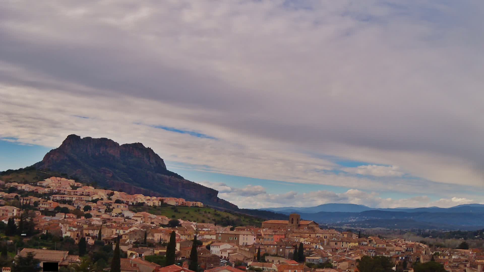 A town of low, tan buildings nestled against a steep, dark mountain under a cloudy sky.