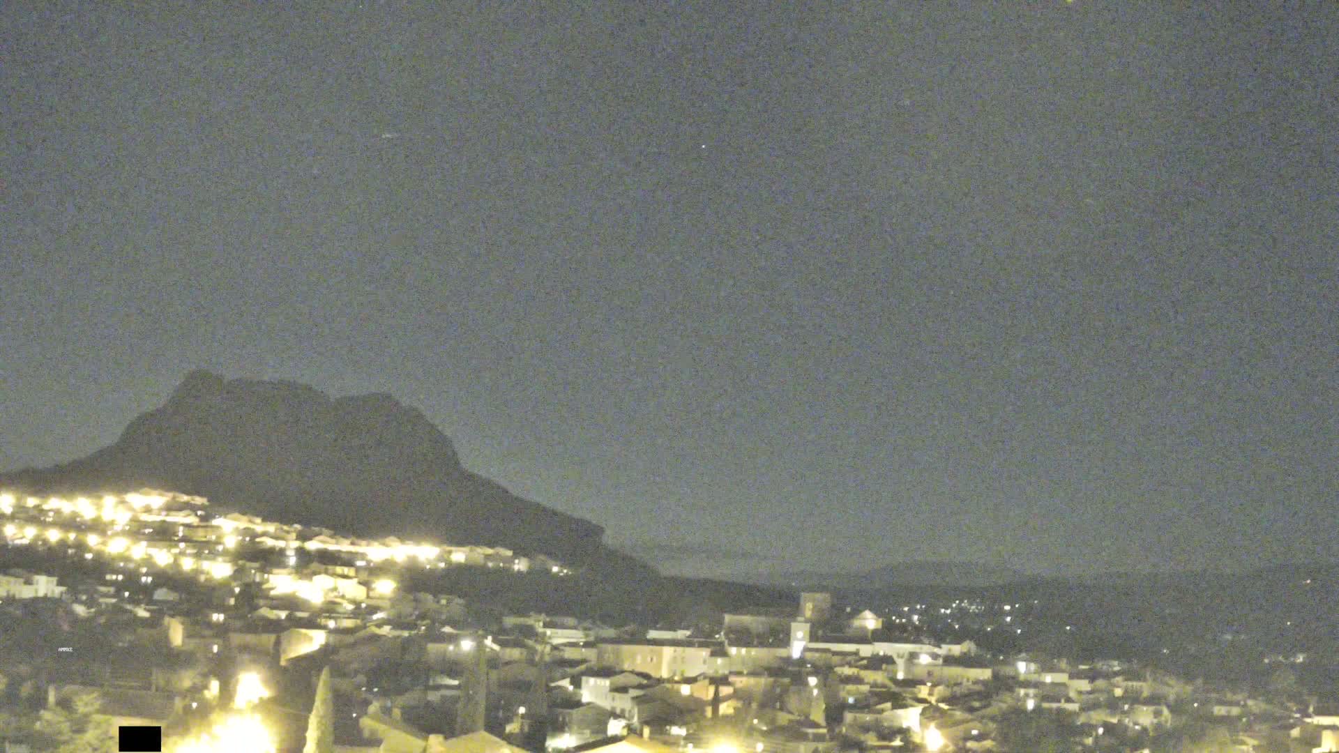 A nighttime view of a town illuminated by lights, nestled beneath a dark mountain under a clear sky.