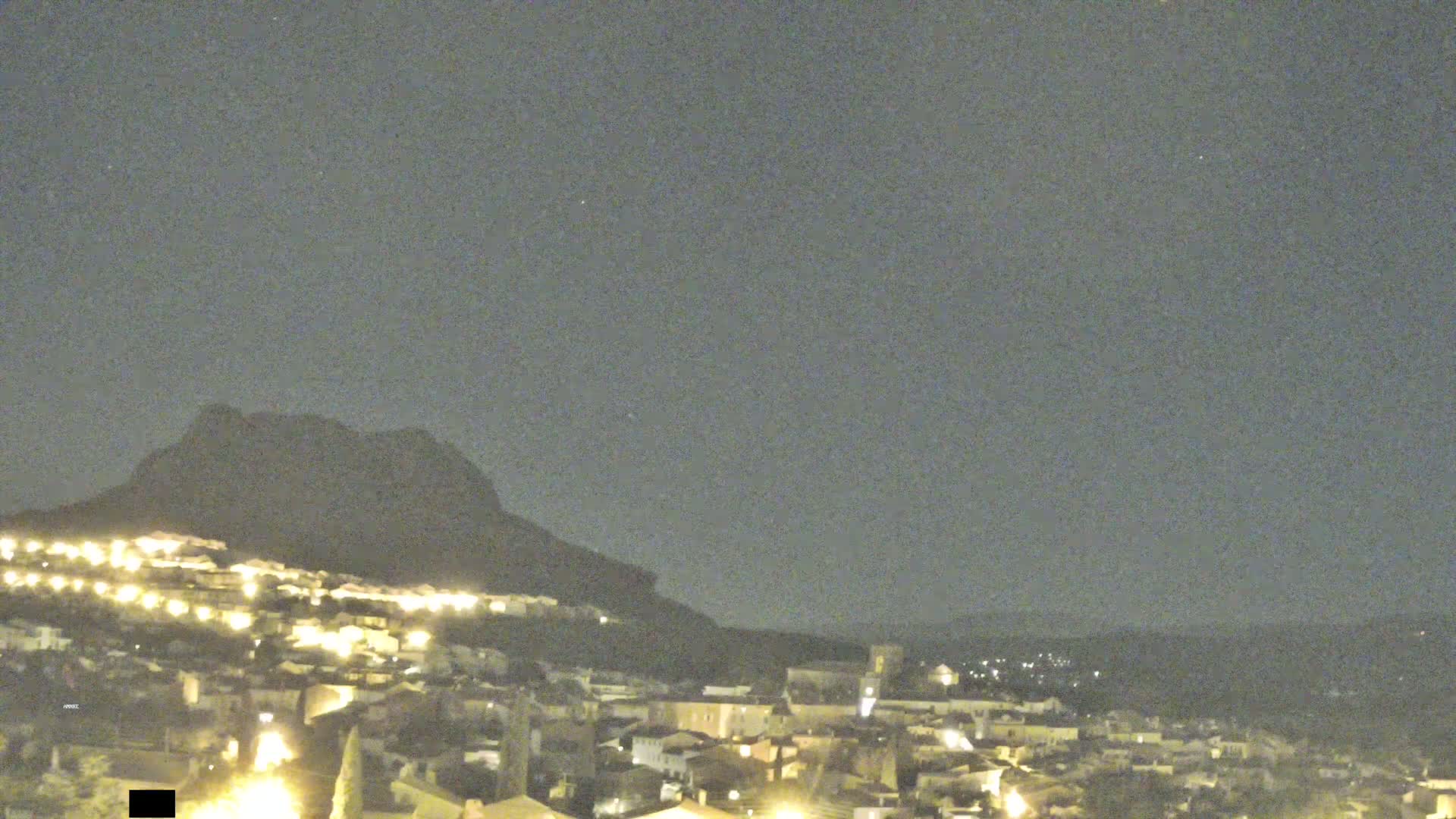 A nighttime view shows a hilltop town illuminated by lights, with a large dark mountain in the background under a clear, dark sky.