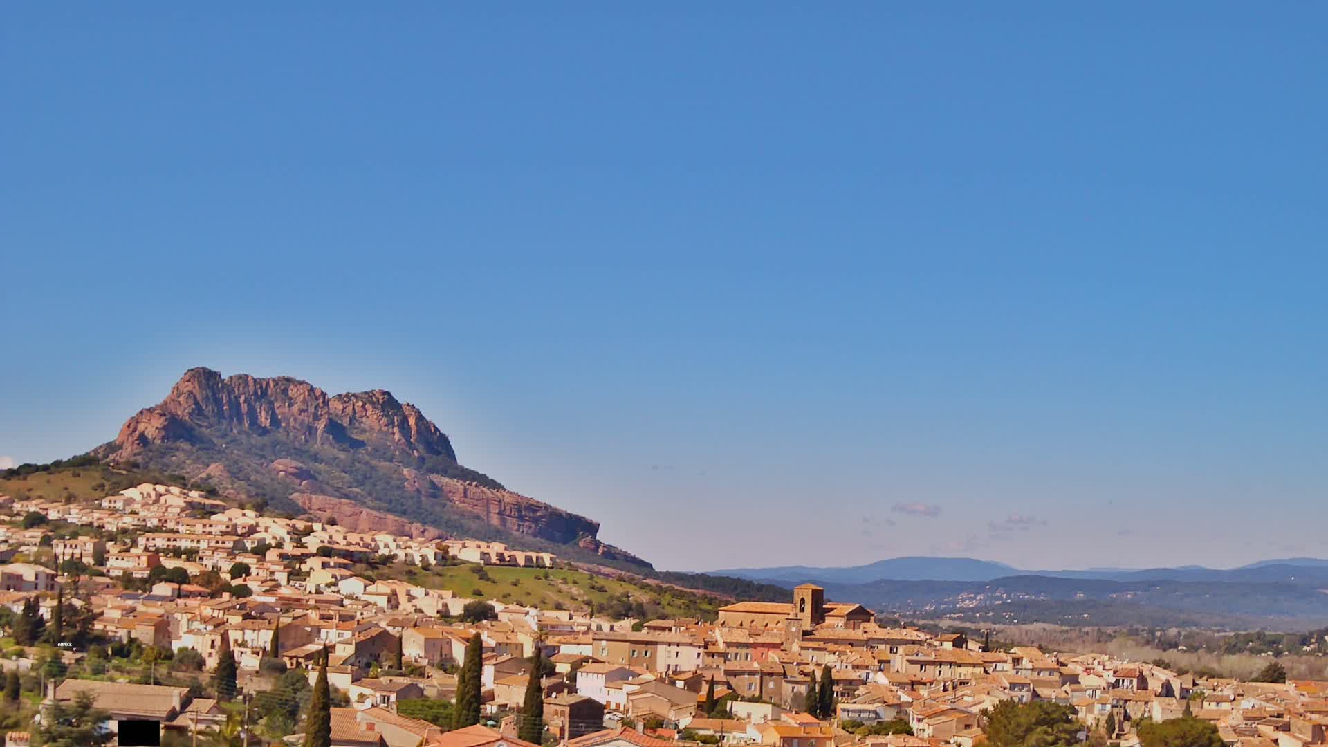 A town of tan and brown buildings sits on a hillside below a large, reddish-brown rock formation under a clear blue sky.