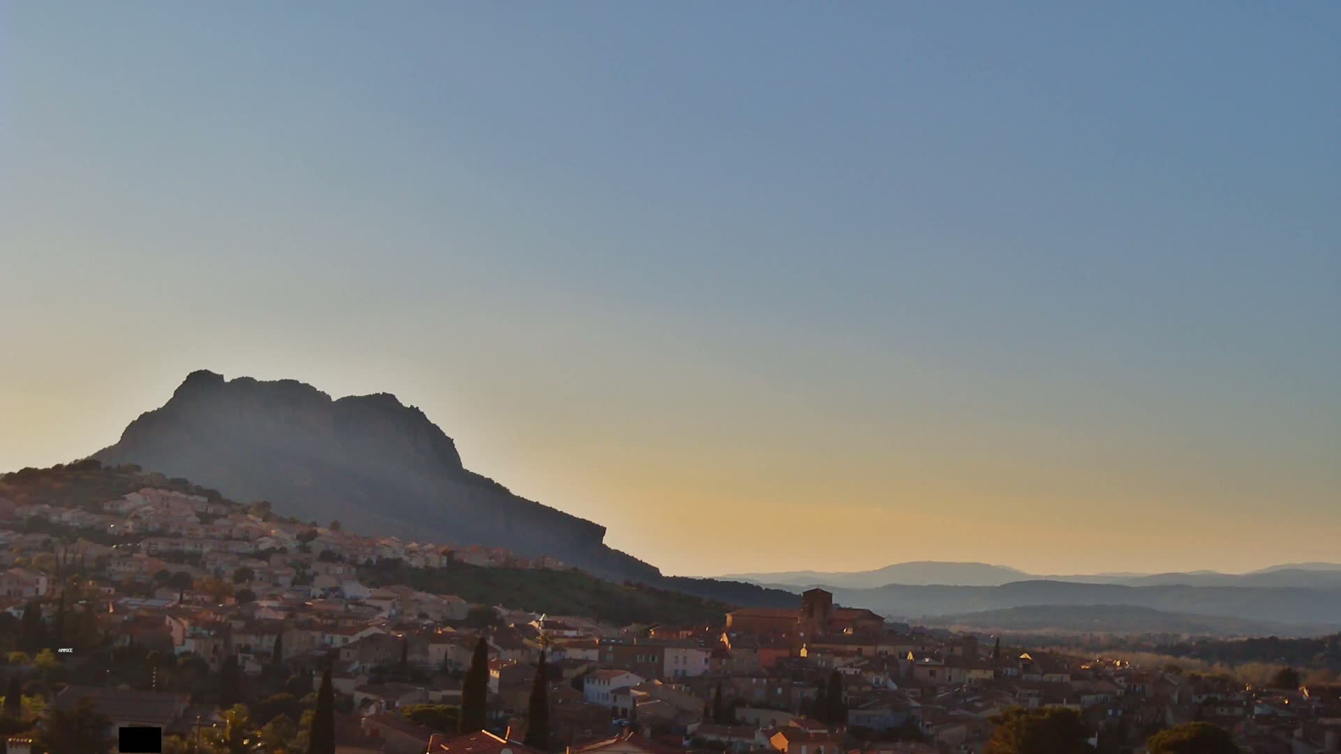 A village nestled at the base of a large, dark mountain, with a hazy sunset illuminating the distant rolling hills.