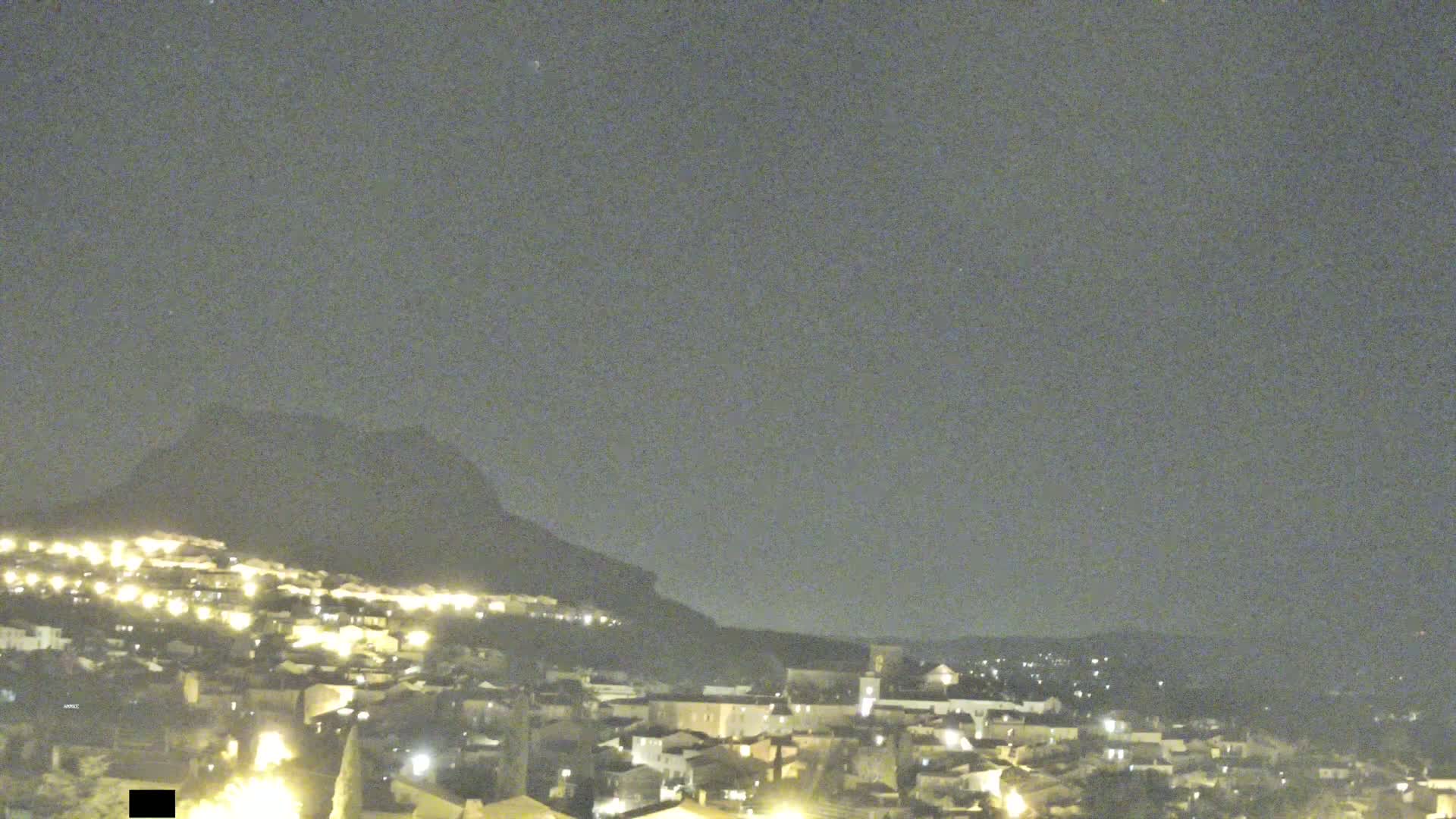 A nighttime view of a small town nestled against a dark hill under a clear, dark sky.