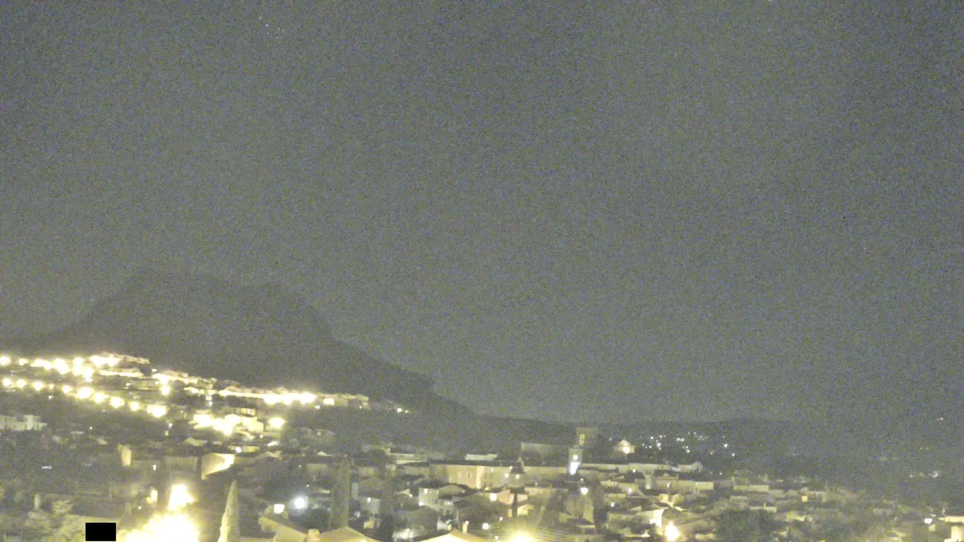A nighttime, overcast view of a small town nestled against a hill, with scattered lights illuminating the buildings.