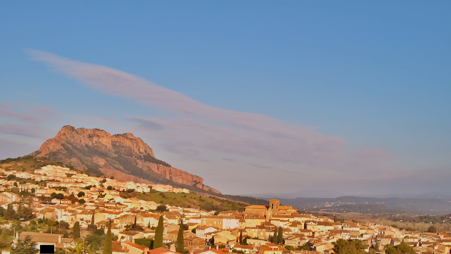 A small town of light-brown buildings nestles against a large, reddish-brown hill under a mostly clear, pale-blue sky with thin, wispy clouds.