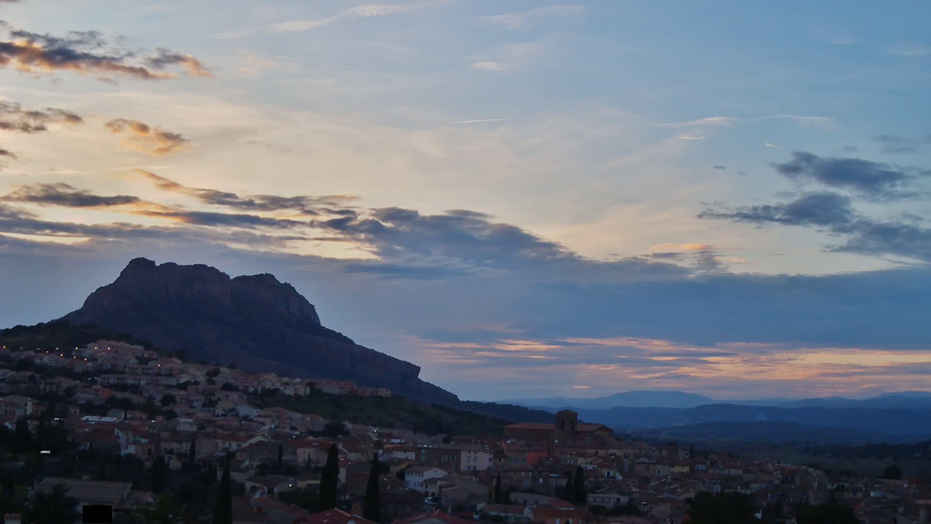 A town nestled at the base of a large, dark mountain is silhouetted against a twilight sky filled with clouds showing hints of pink and orange.