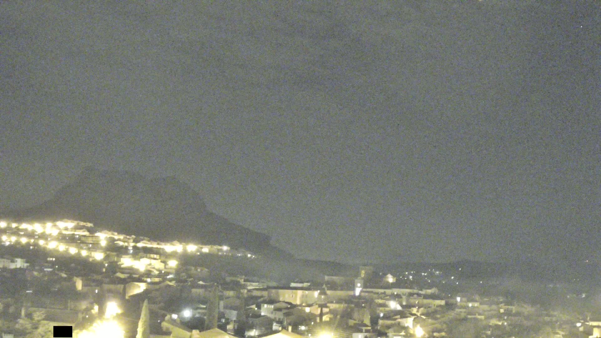 A nighttime view of a small town nestled against a dark hill under a cloudy sky.