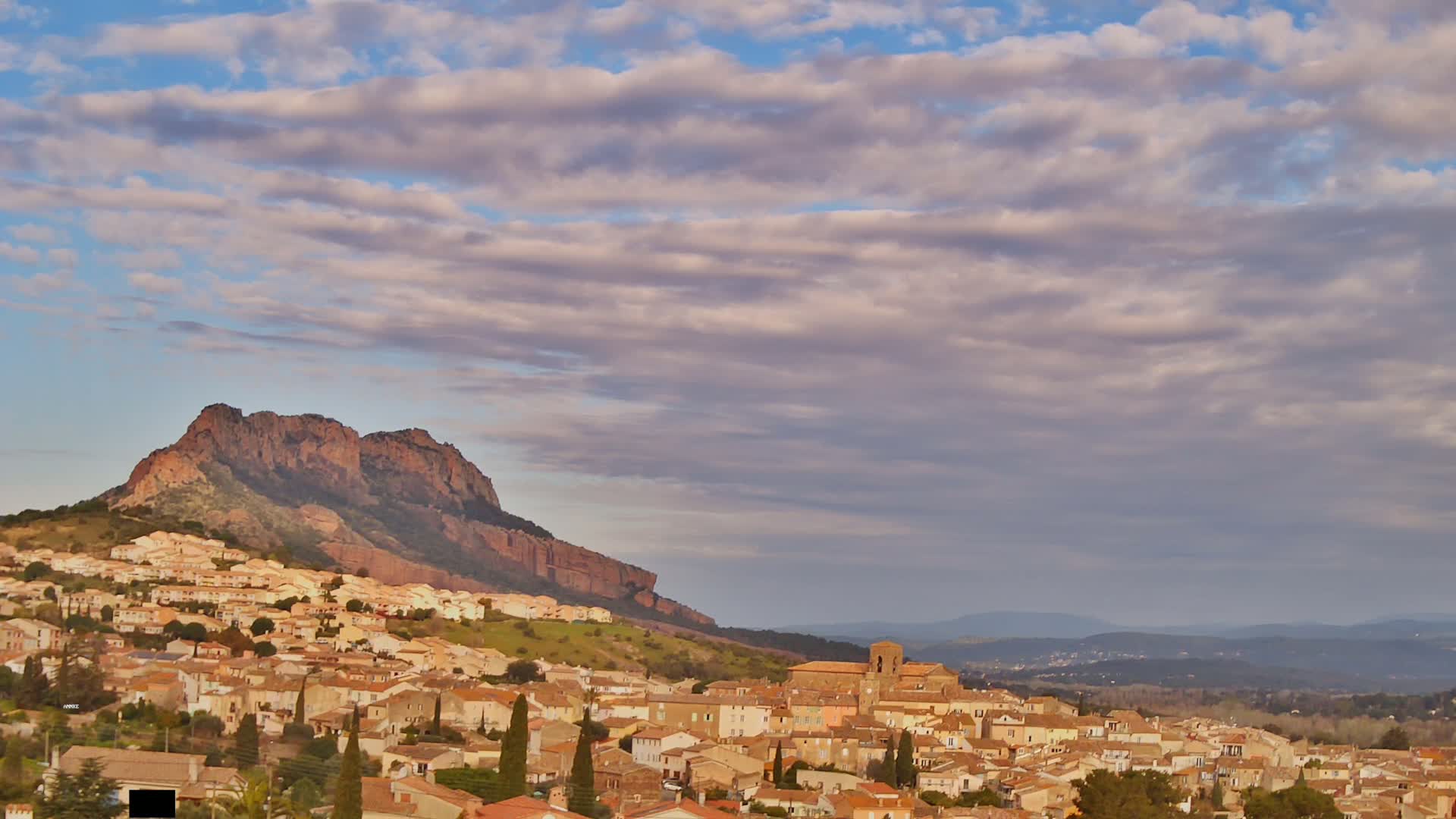 A hillside town with tan-colored buildings is nestled below a large, reddish-brown rocky mountain under a partly cloudy sky.