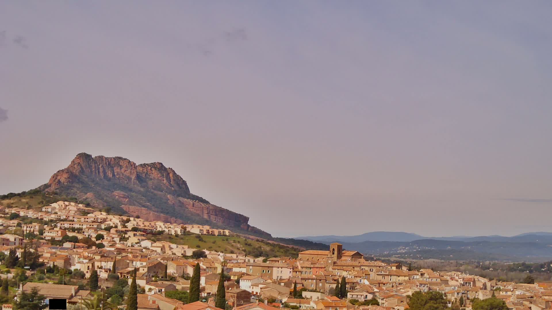 A hilltop village of tan and brown buildings clusters beneath a large, rocky mountain under a pale, hazy sky.