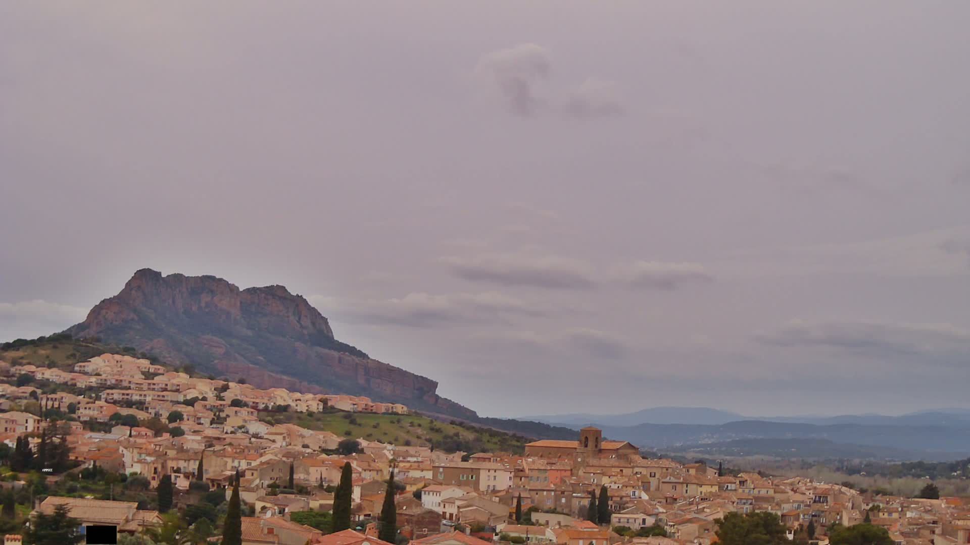A hilltop town of tan buildings stretches across a hillside beneath a large, dark-colored mountain under a pale overcast sky.