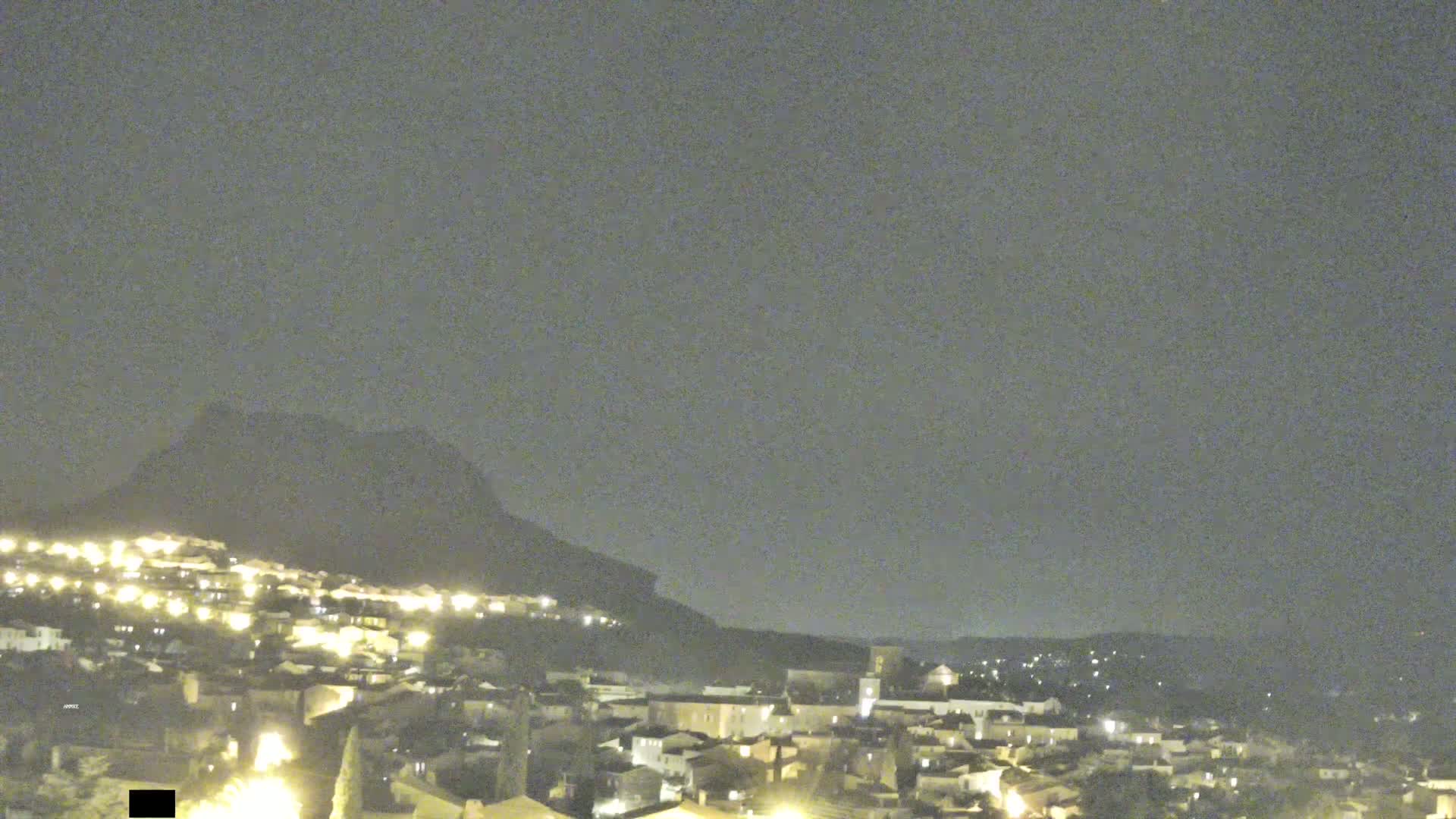 A nighttime view of a town nestled against a dark mountain under a cloudy sky.