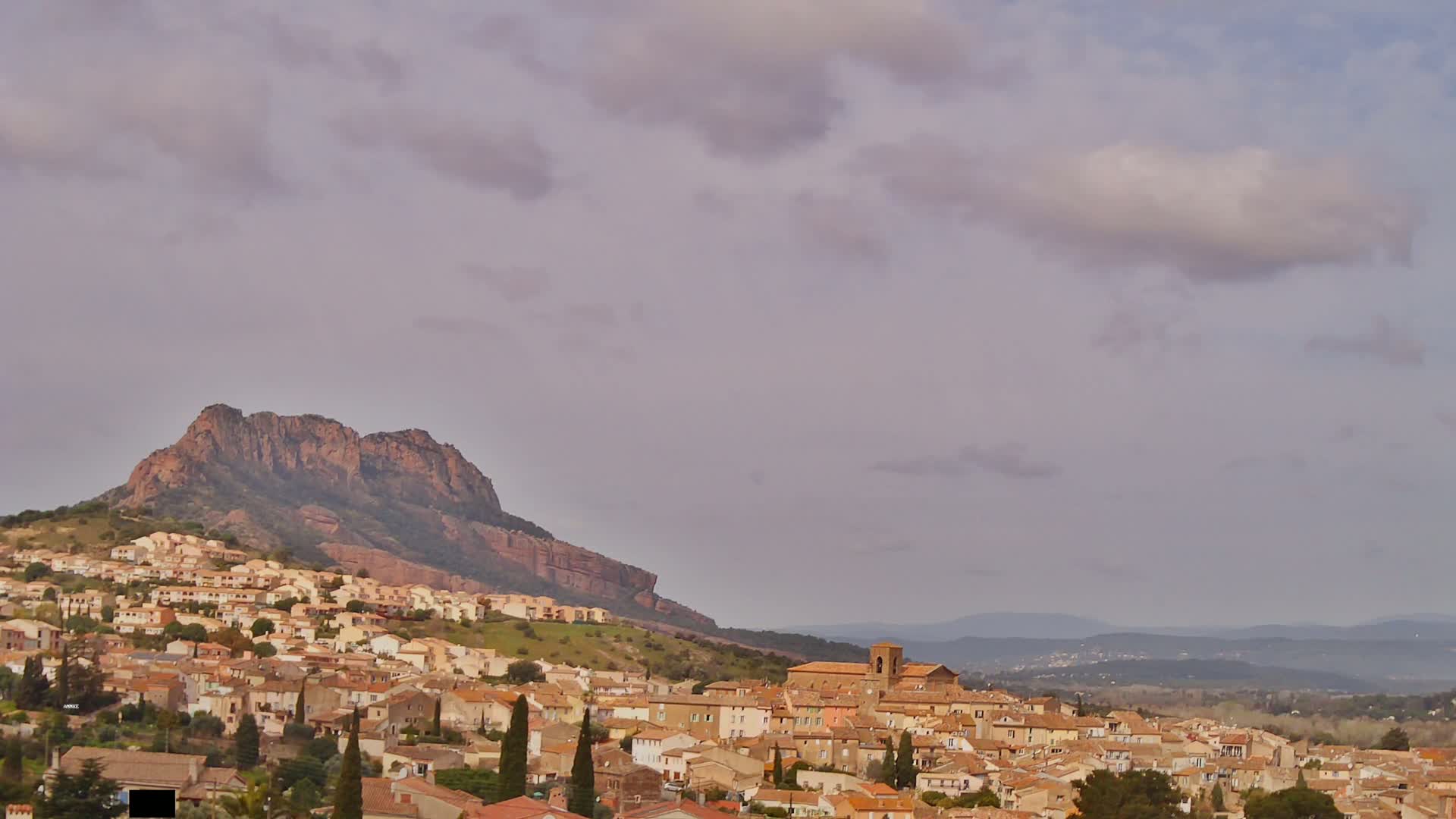 A hilly town with tan-colored buildings is nestled beneath a large, reddish-brown mountain under a mostly cloudy sky.
