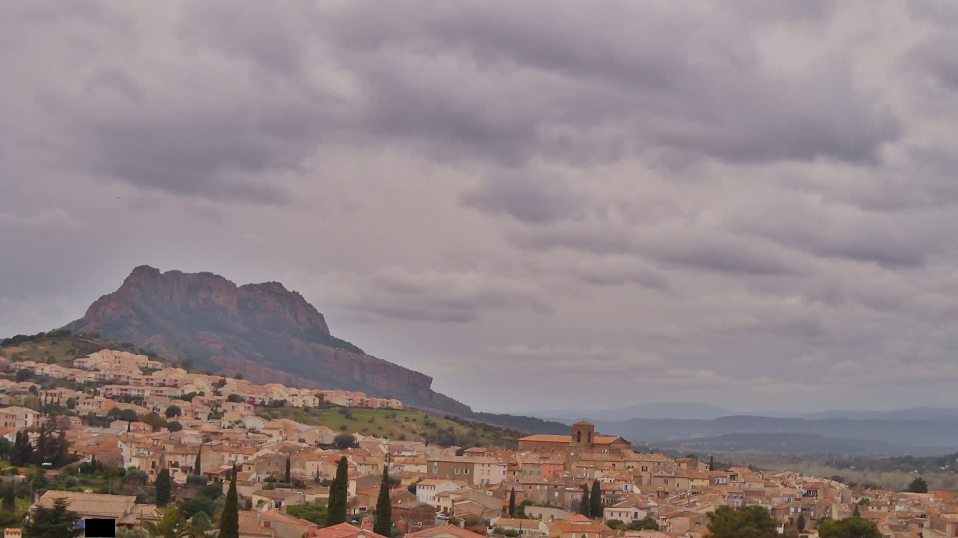A hilly town of tan buildings nestles beneath a large, dark-colored mountain under a cloudy sky.