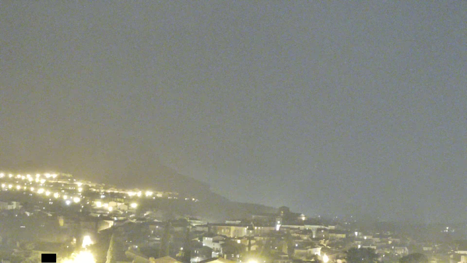 A hazy night scene shows a town illuminated with scattered lights, nestled against a hill under a muted, overcast sky.