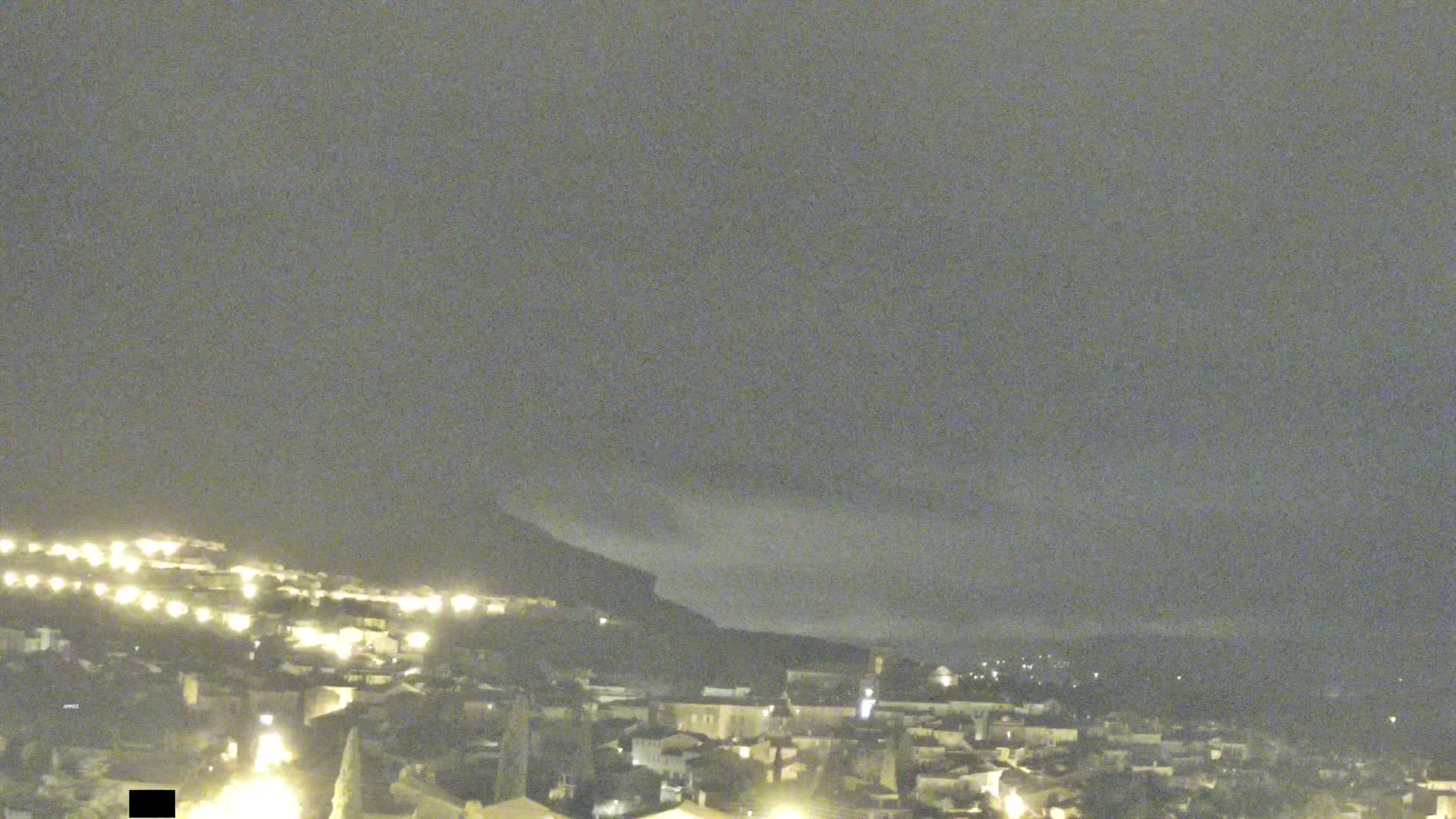 A nighttime view of a town nestled against a hill, under a cloudy sky.
