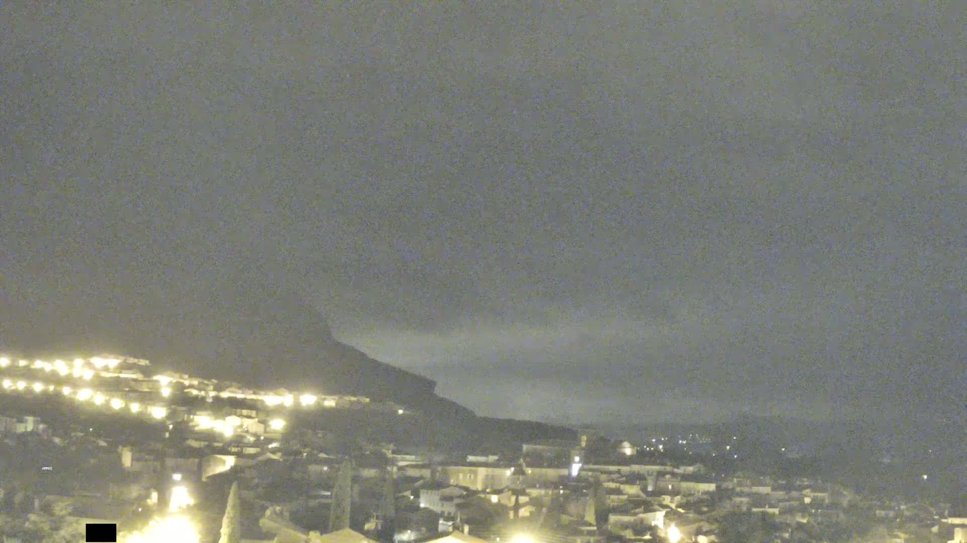 A nighttime view of a town nestled at the base of a hill, under a mostly cloudy sky.