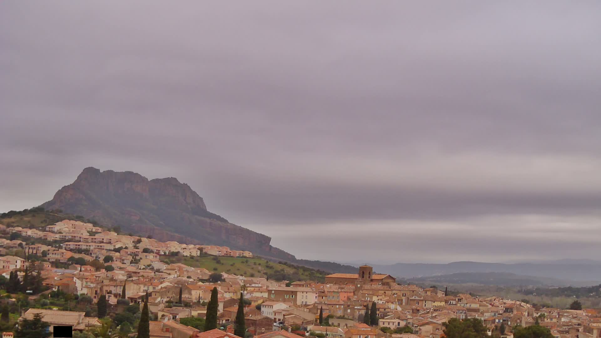 A hilltop town with tan-colored buildings stretches across a hillside beneath an overcast sky, backed by a large, dark mountain.