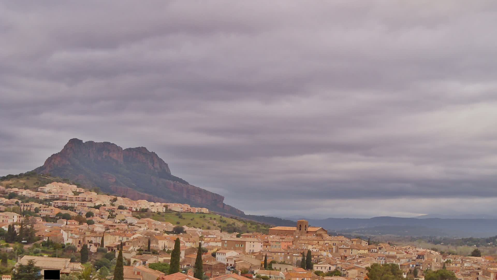 A large hill with a village of tan buildings on its slopes is seen under a cloudy sky.
