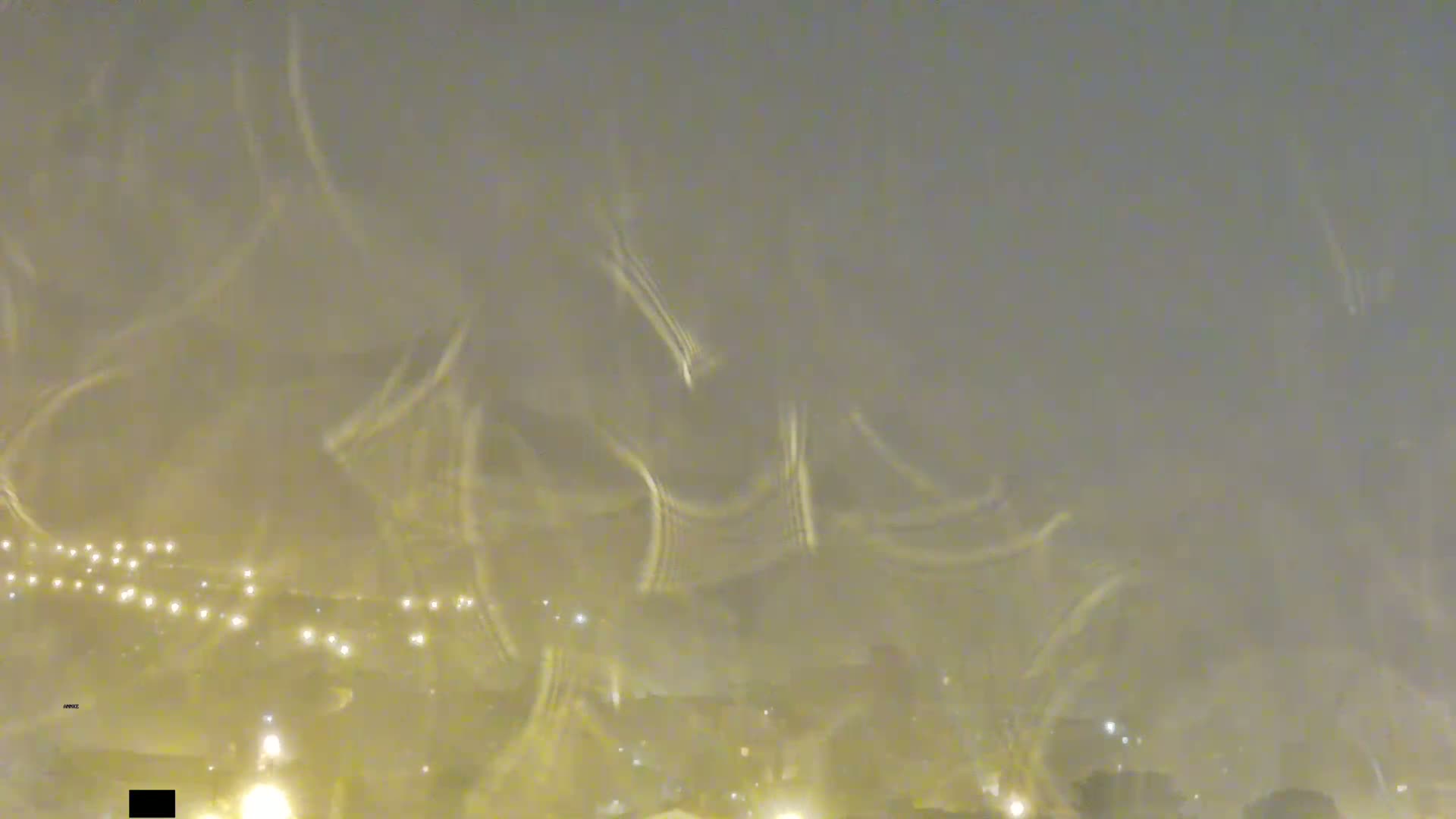 A blurry nighttime view of a snow-covered landscape with distant lights visible through falling snow.